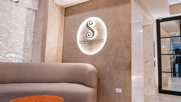 Un moderno vestíbulo de hotel con un sofá mullido, una mesa de centro de madera y un logotipo iluminado empotrado en la pared para Hotel & Residences Cebu.