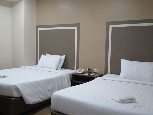 Habitación de hotel con dos camas individuales, cada una con ropa blanca y almohadas. Hay una mesita de noche de madera con un teléfono y una pequeña mesa con un cartel y un enchufe.