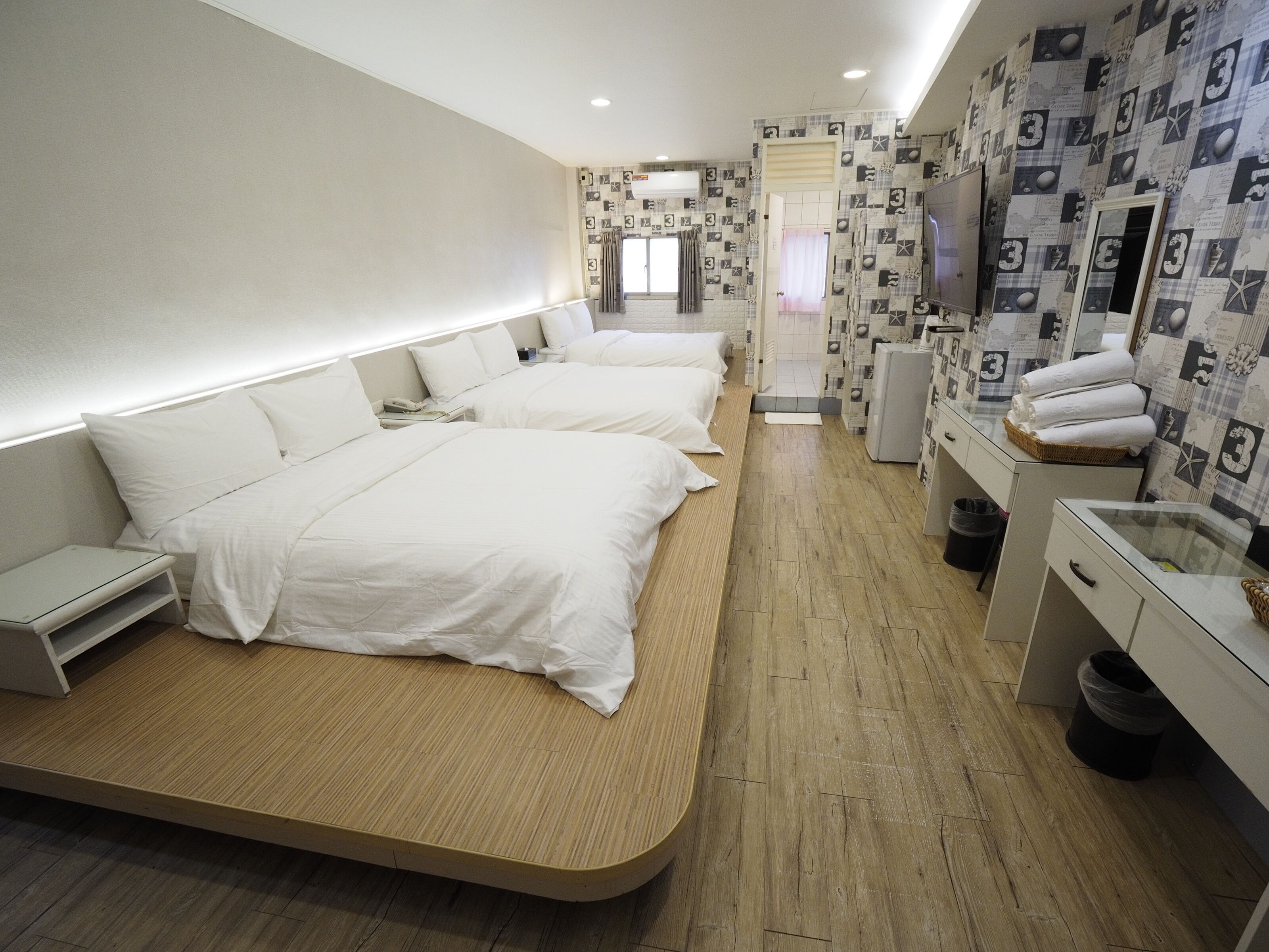 A modern hotel room with three beds, a small desk, a TV, and a bathroom with a mirror and towel rack.