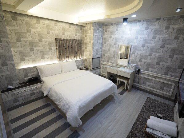 A modern hotel room with a large bed, bedside table with a lamp, a vanity with a mirror, and a small stool.