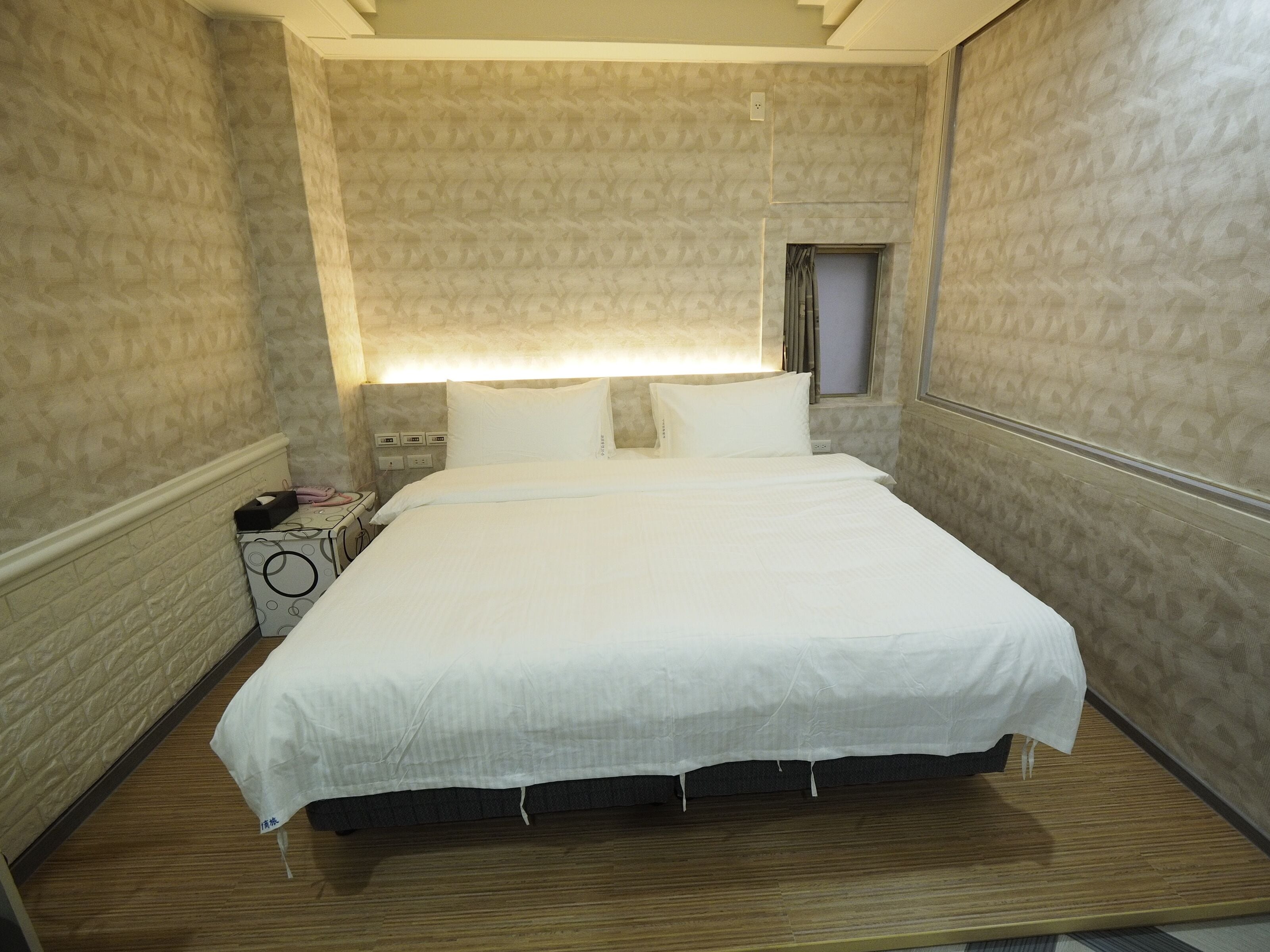 A hotel room with a large bed, a bedside table with a lamp, and a small window.