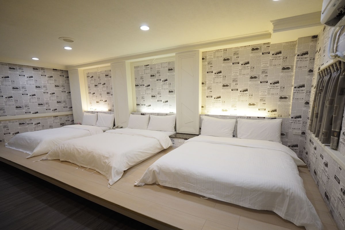 A hotel room with four single beds arranged in a row, each with white bedding and pillows. The walls are covered with a newspaper print wallpaper.
