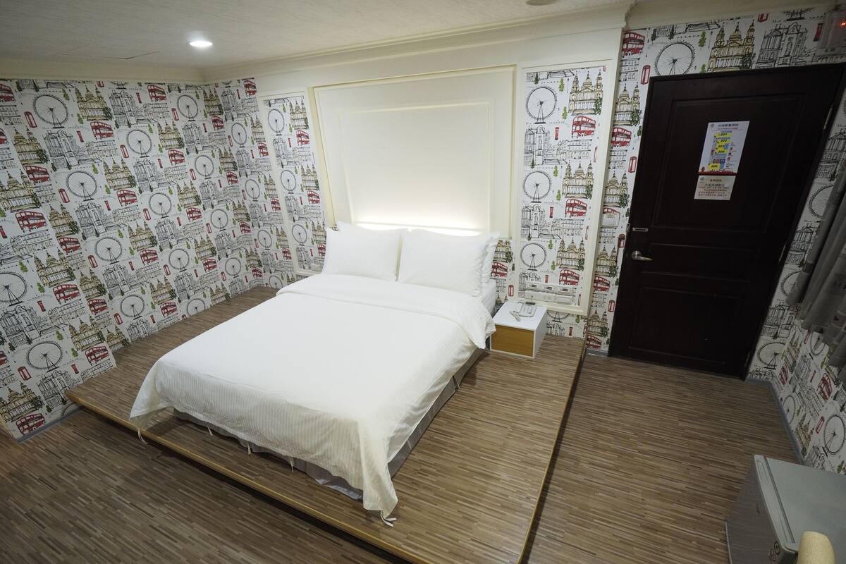 A hotel room with a bed, wooden flooring, a nightstand, a small table, and a door.
