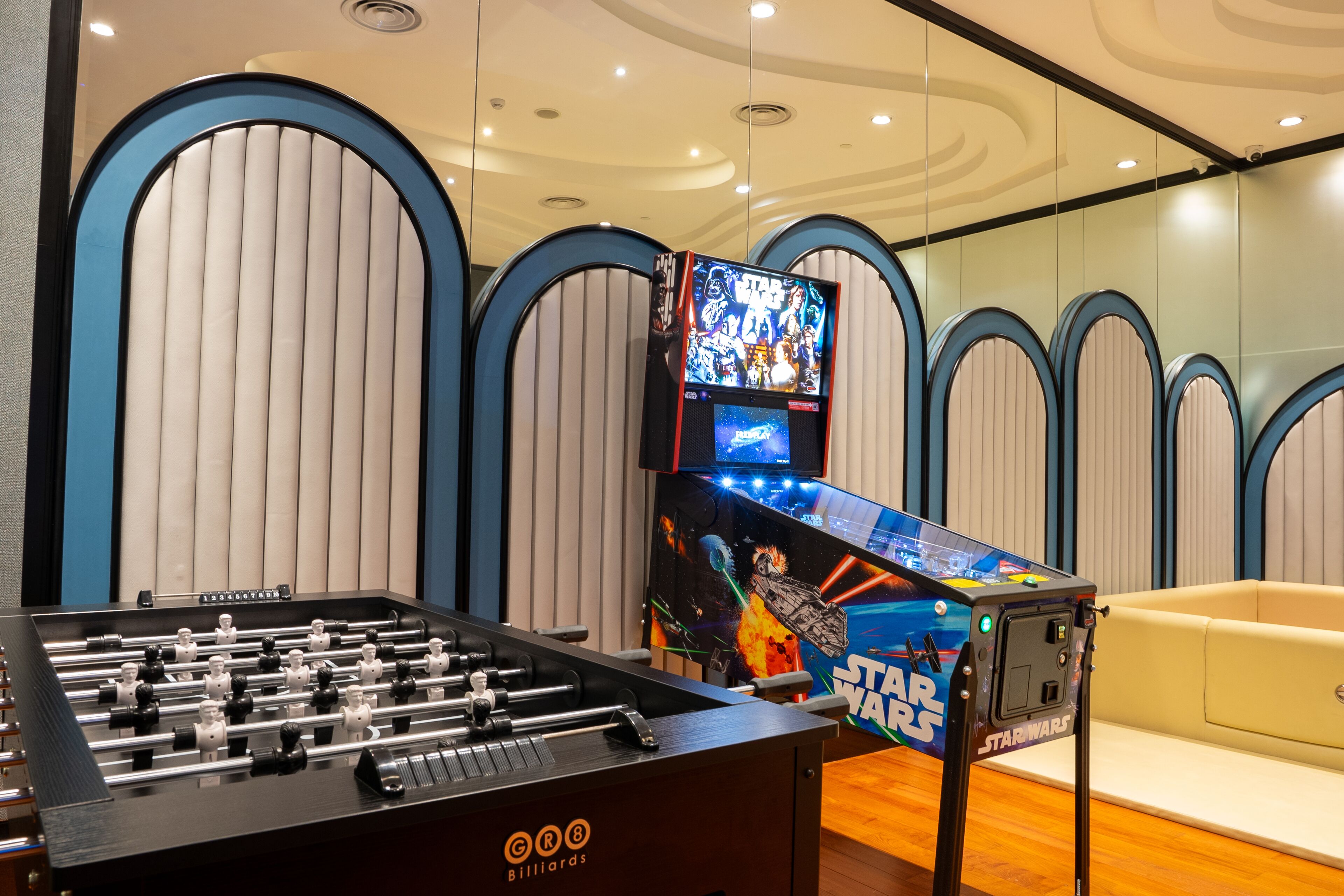 A foosball table and a Star Wars arcade game in a room with a beige sofa and a modern ceiling design.