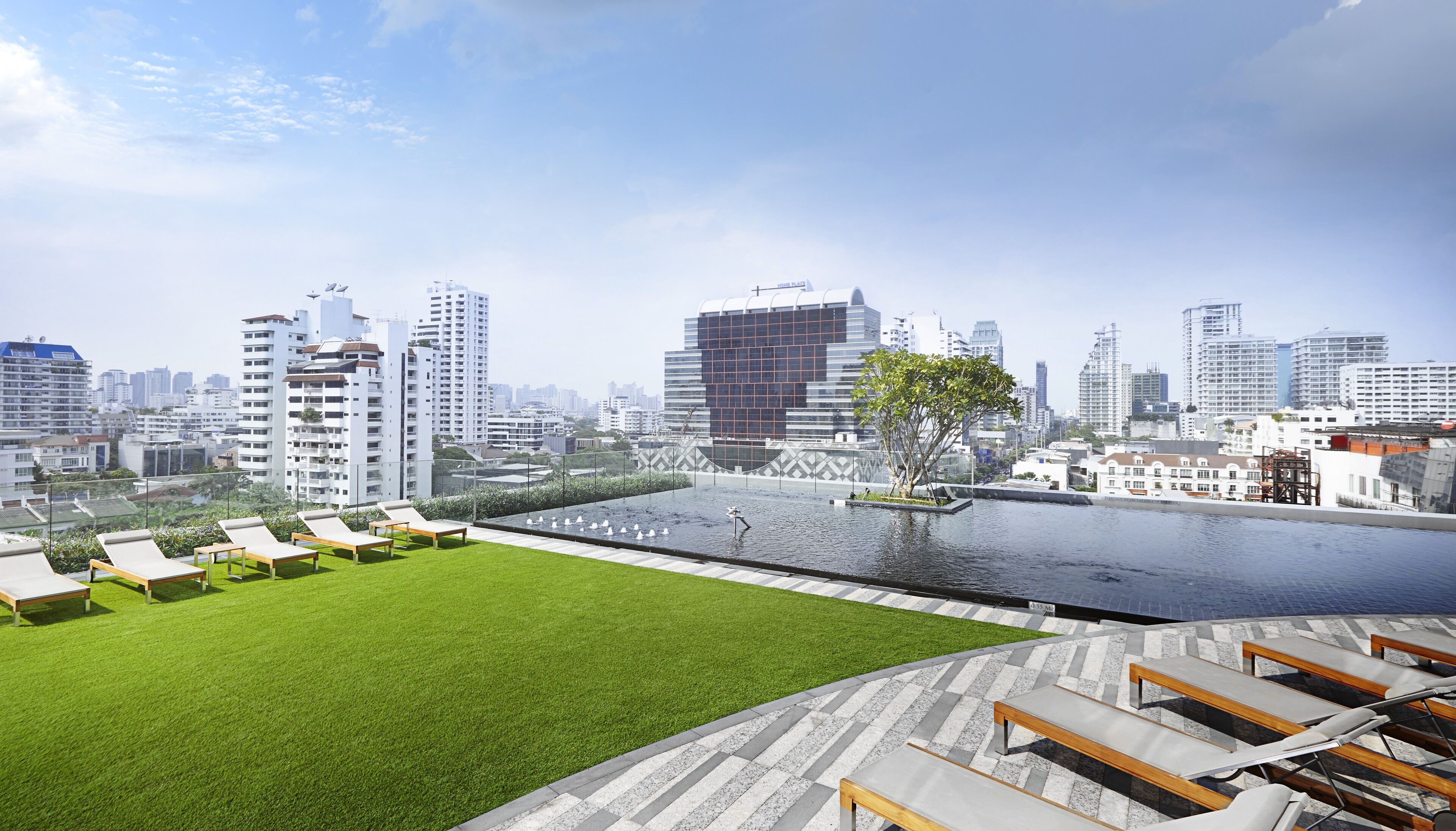 A rooftop pool area with lounge chairs, a small water feature, and a view of a cityscape with modern buildings.