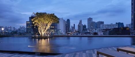 Un paisaje urbano frente al agua con edificios modernos, un árbol iluminado por luces y una masa de agua tranquila.