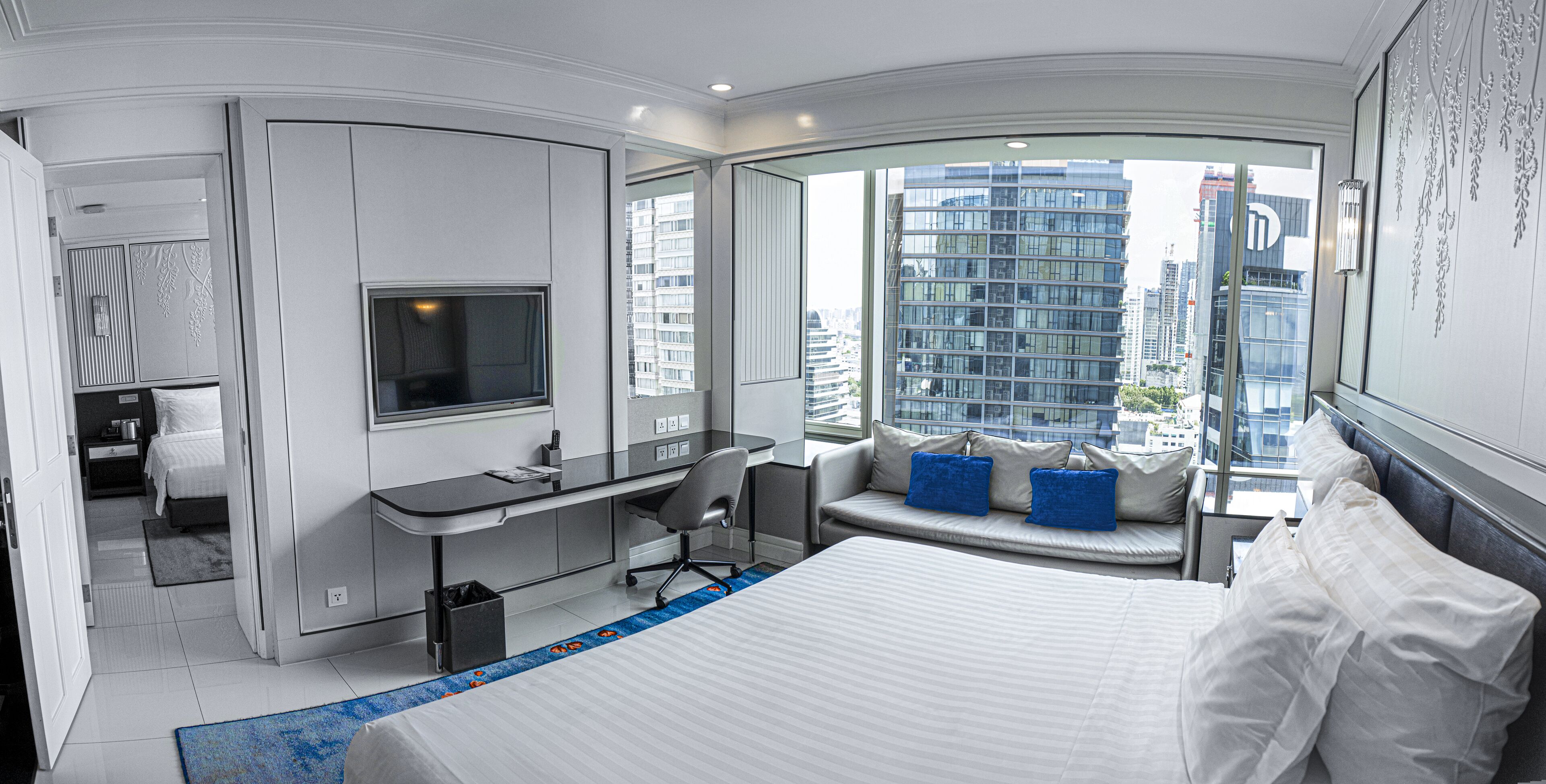 Habitación de hotel moderna con una cama grande, un escritorio con silla, televisión y vistas de la ciudad a través de amplias ventanas.