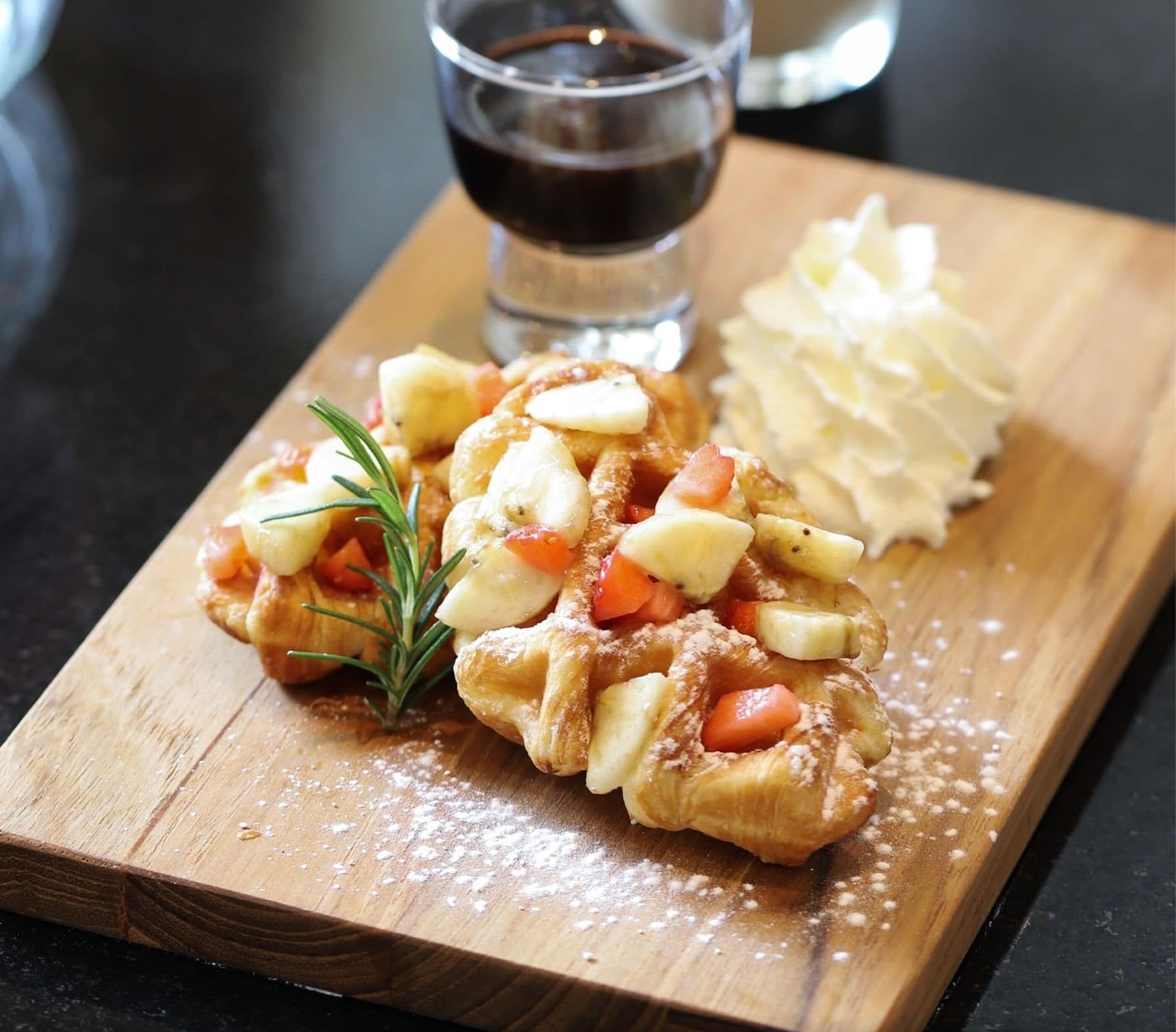 A wooden board with a pastry topped with fruit and a sprig of rosemary, accompanied by a glass of dark liquid and a side of whipped cream.