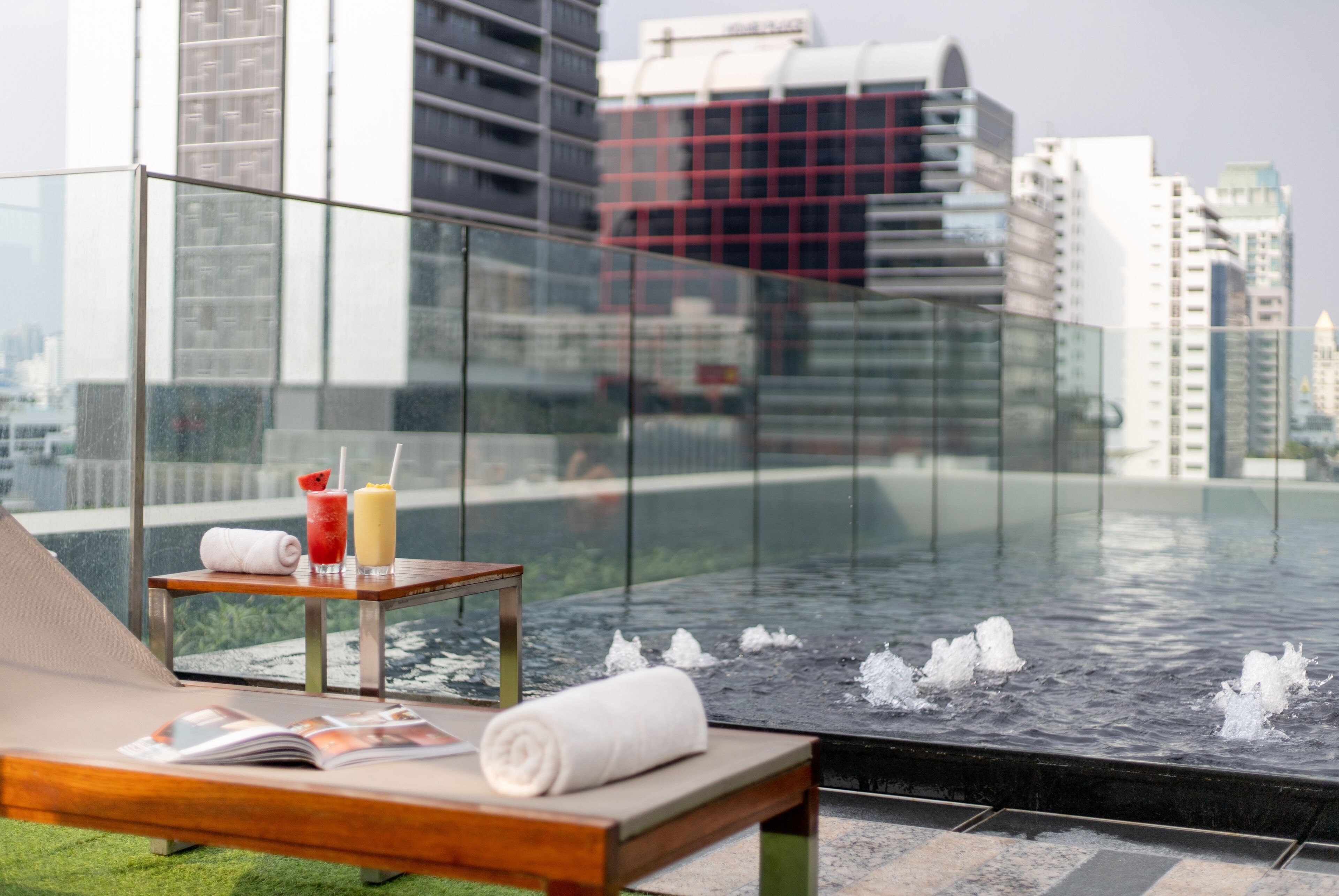Un lounge en la azotea con barandilla de cristal, una piscina con hidromasajes y vistas a edificios de la ciudad.