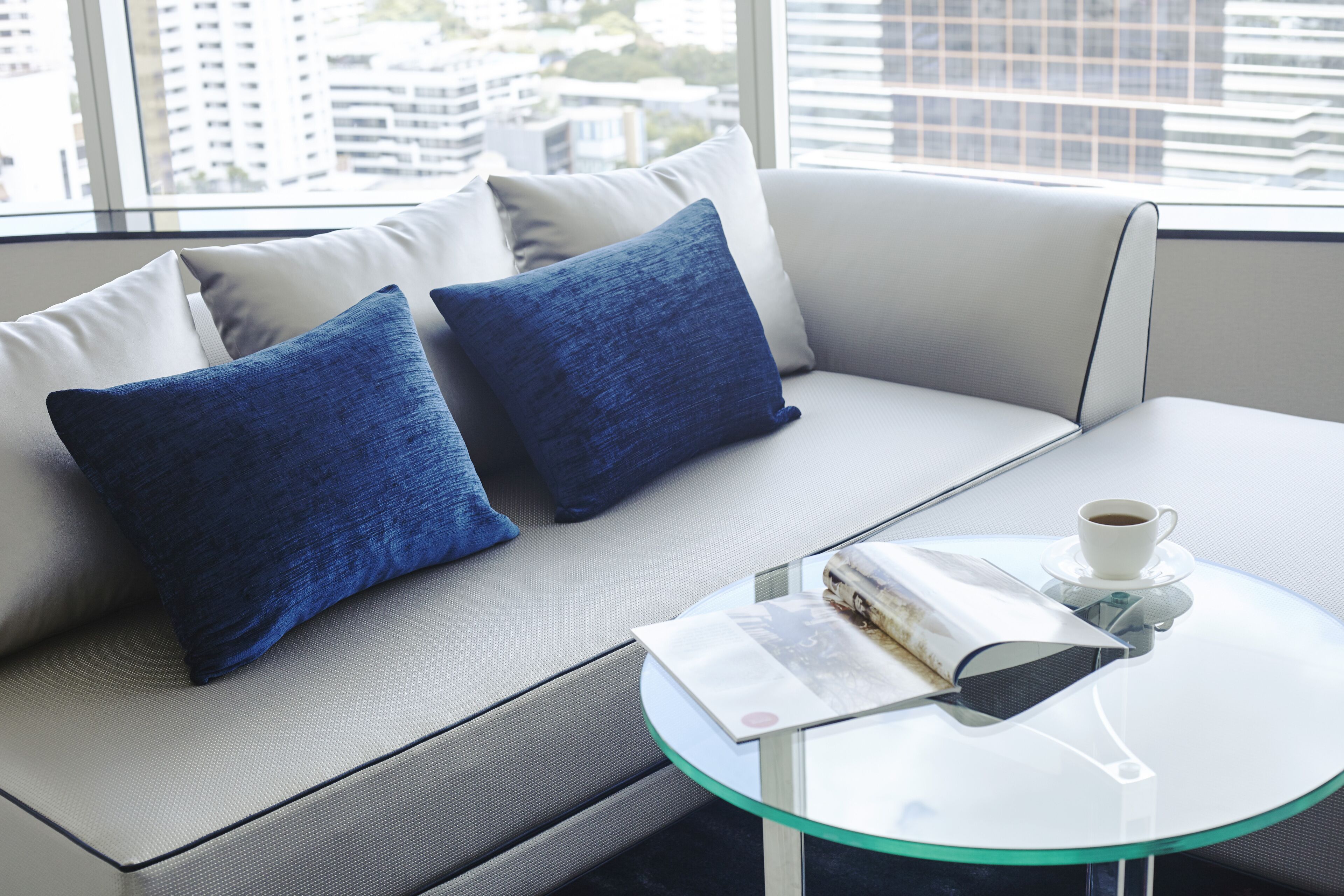 A modern living room with a sofa, two blue cushions, a glass coffee table with a magazine and a cup of coffee.