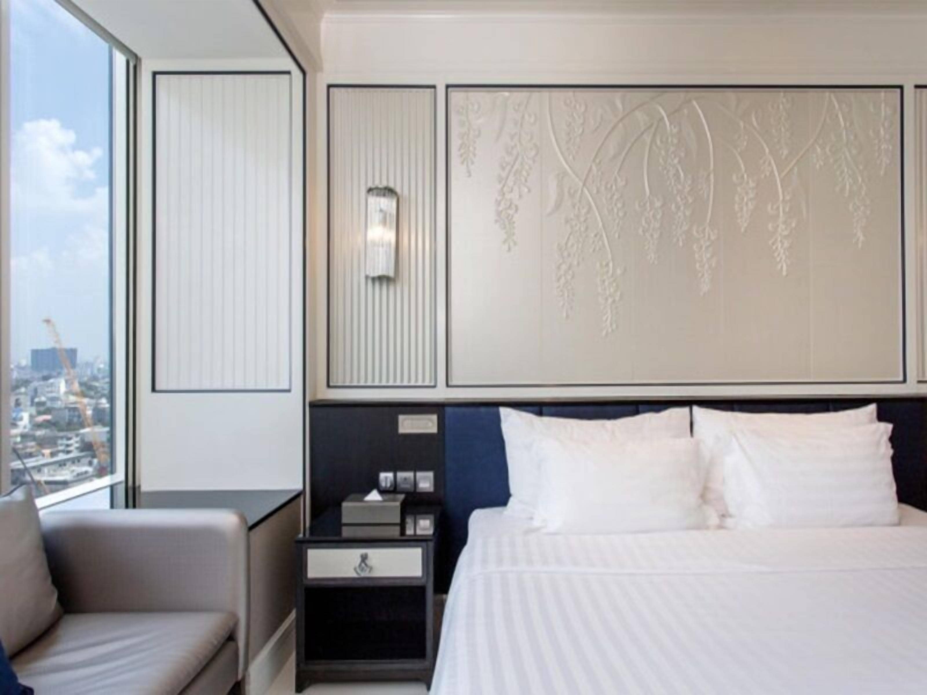A hotel room with a large bed, a nightstand, a chair, and a view of the city.