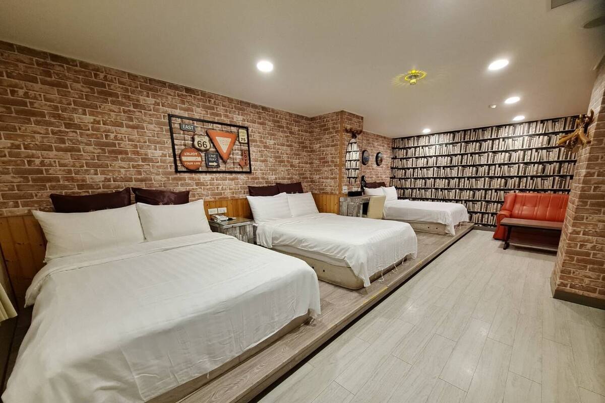 Une chambre d’hôtel avec trois lits, un mur en briques, une bibliothèque et un canapé en cuir rouge.
