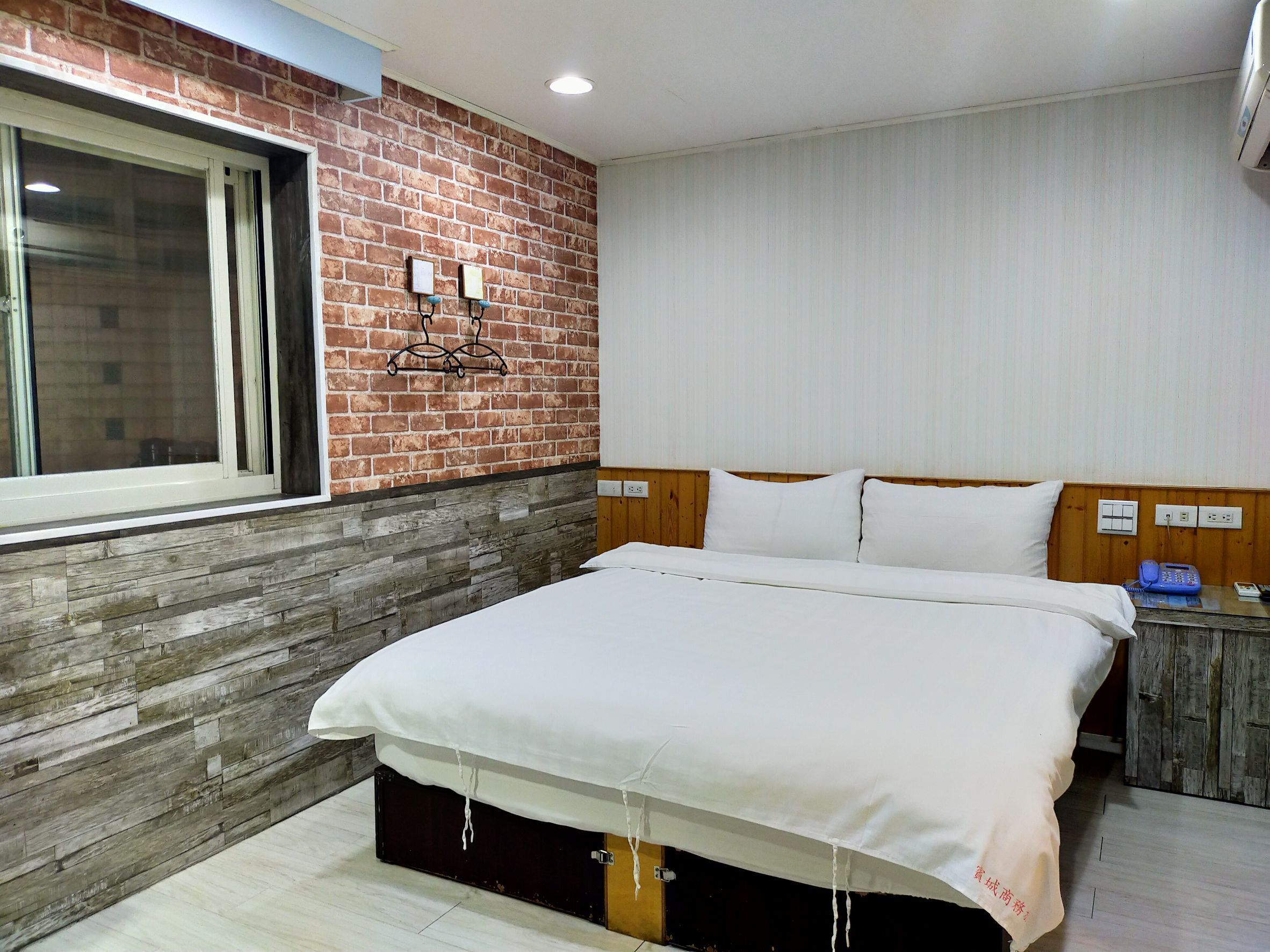 Habitación de hotel con pared de ladrillo decorativa, cabecera de madera, cama grande con ropa blanca, mesita de noche con teléfono y ventana con persianas.