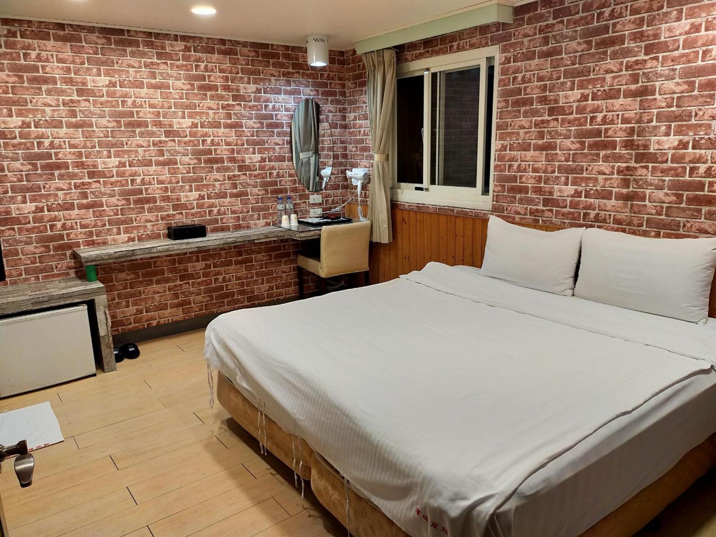 A hotel room with a brick wall, a wooden bed, a desk with a chair, a window, and a mirror.