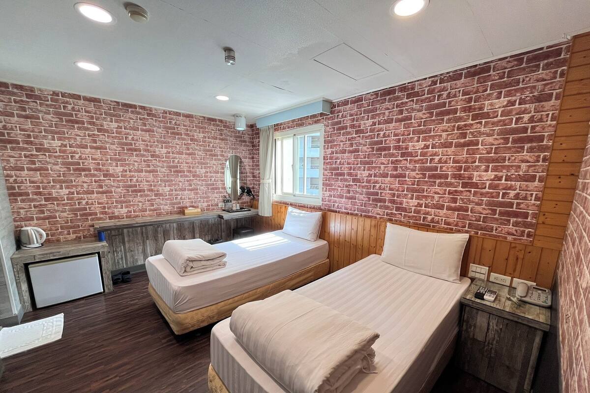 Une chambre d’hôtel avec un mur en briques, deux lits, un petit bureau et une fenêtre.