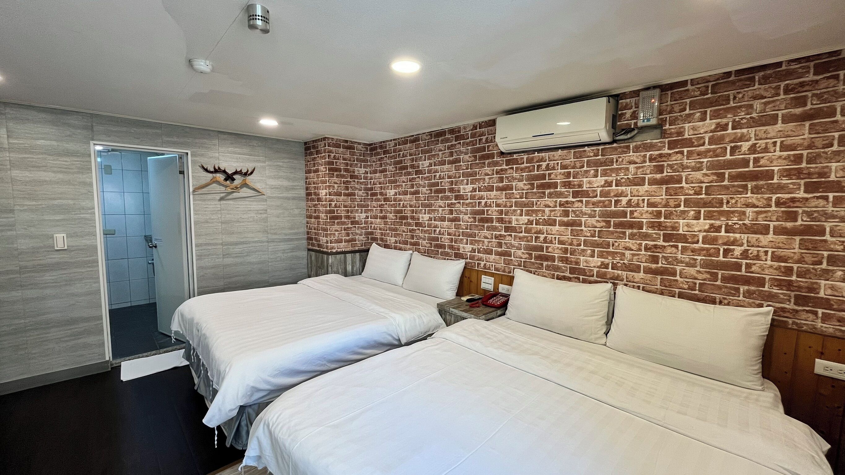 Habitación de hotel con dos camas, una pared de ladrillo, un equipo de aire acondicionado y una puerta que da al baño.
