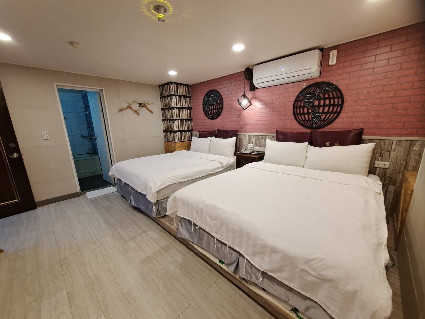 A hotel room with two beds, a bookshelf, a wall-mounted air conditioner, and a door leading to a bathroom.