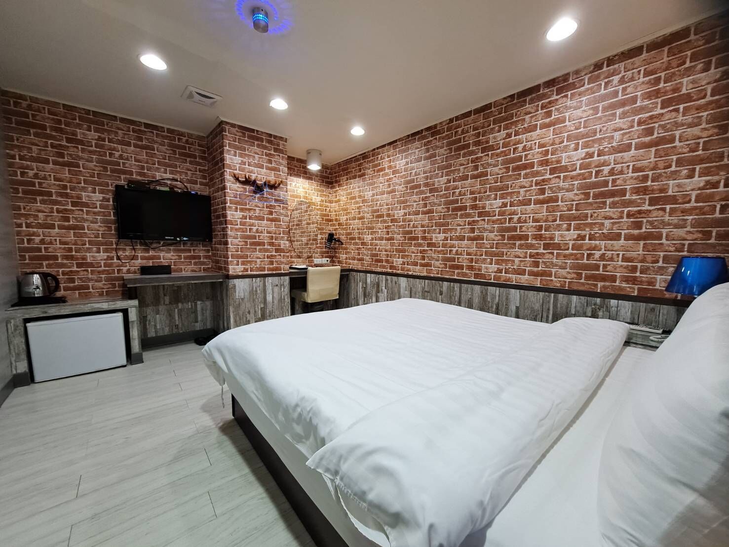Habitación de hotel con pared de ladrillo, televisor de pantalla plana montado en la pared, una mesita de noche con una tetera, cama con ropa blanca y una lámpara azul en la mesita de noche.