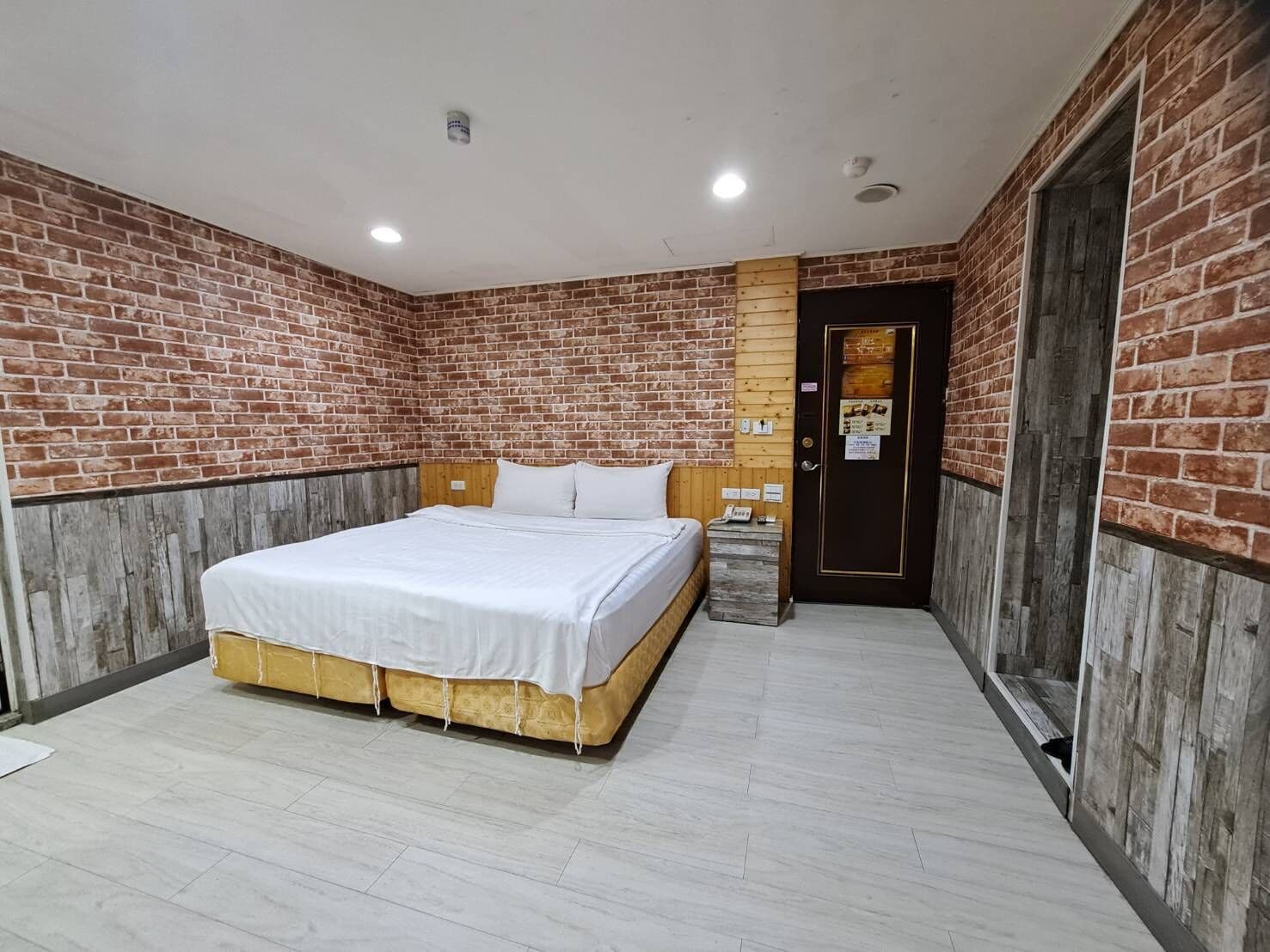 A hotel room with a brick wall, a wooden headboard, a bed with white bedding, a nightstand, and a door with a sign.