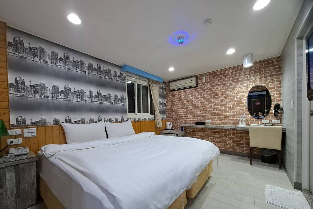 Une chambre avec un lit, une fresque représentant un paysage urbain, un mur en briques, un miroir et une petite kitchenette.
