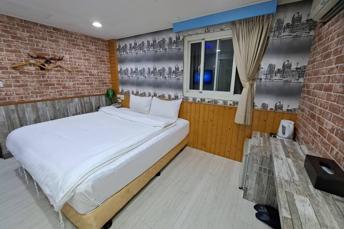 Une chambre avec un lit, une fresque représentant un paysage urbain, une fenêtre avec des rideaux et un mur en briques.