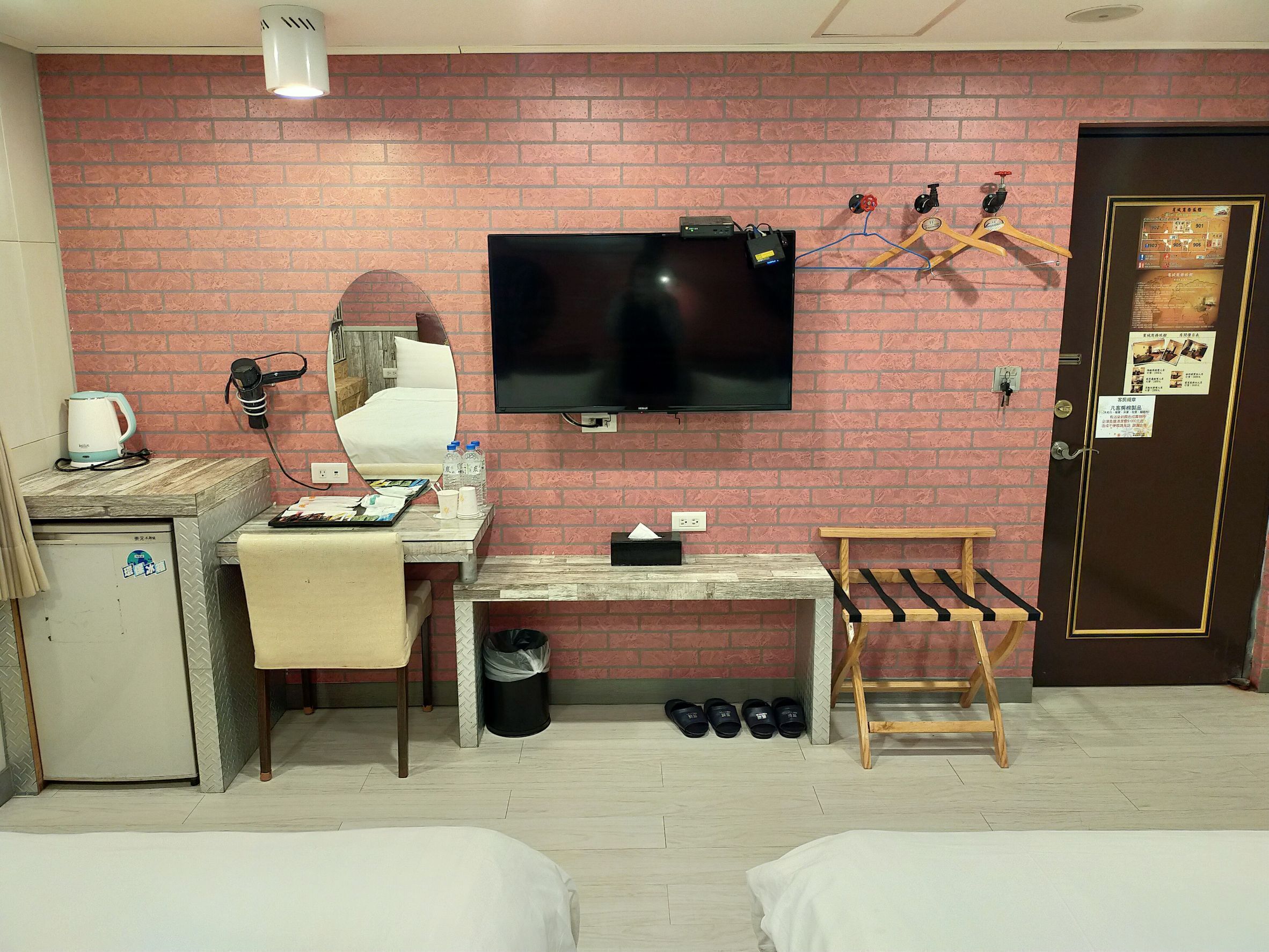 Una camera d'albergo compatta con parete a motivi a mattoni, TV a schermo piatto, scrivania con bollitore, sedia, letto, un piccolo tavolo e un porta scarpe.