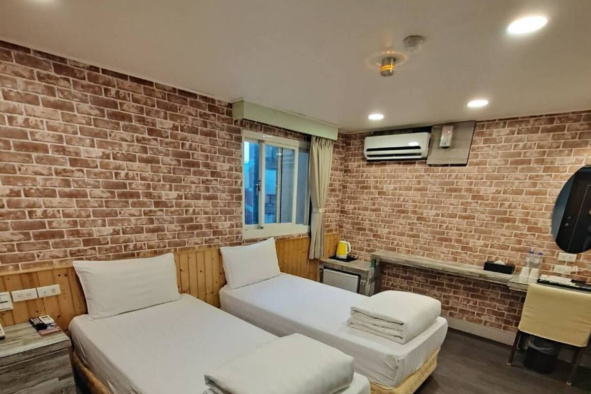 Une chambre d’hôtel avec deux lits, une tête de lit en bois, un mur en briques, une fenêtre avec des rideaux, un appareil de climatisation, une table de chevet et un petit bureau avec une chaise.