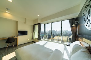 Habitación de hotel con una cama grande, un escritorio, una silla, un televisor y vistas de la ciudad a través de amplios ventanales.