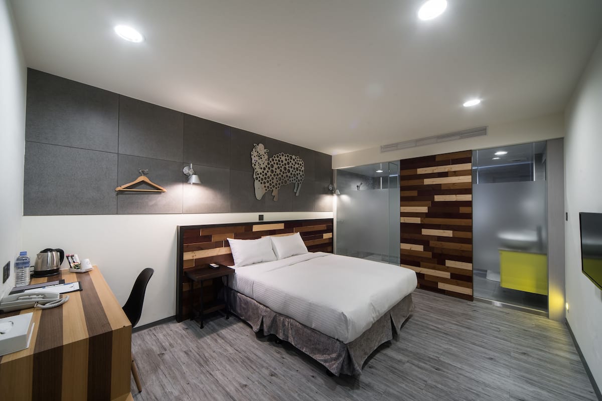 Une chambre d’hôtel moderne avec une tête de lit en bois, un bureau avec une chaise, un lit et un grand miroir.