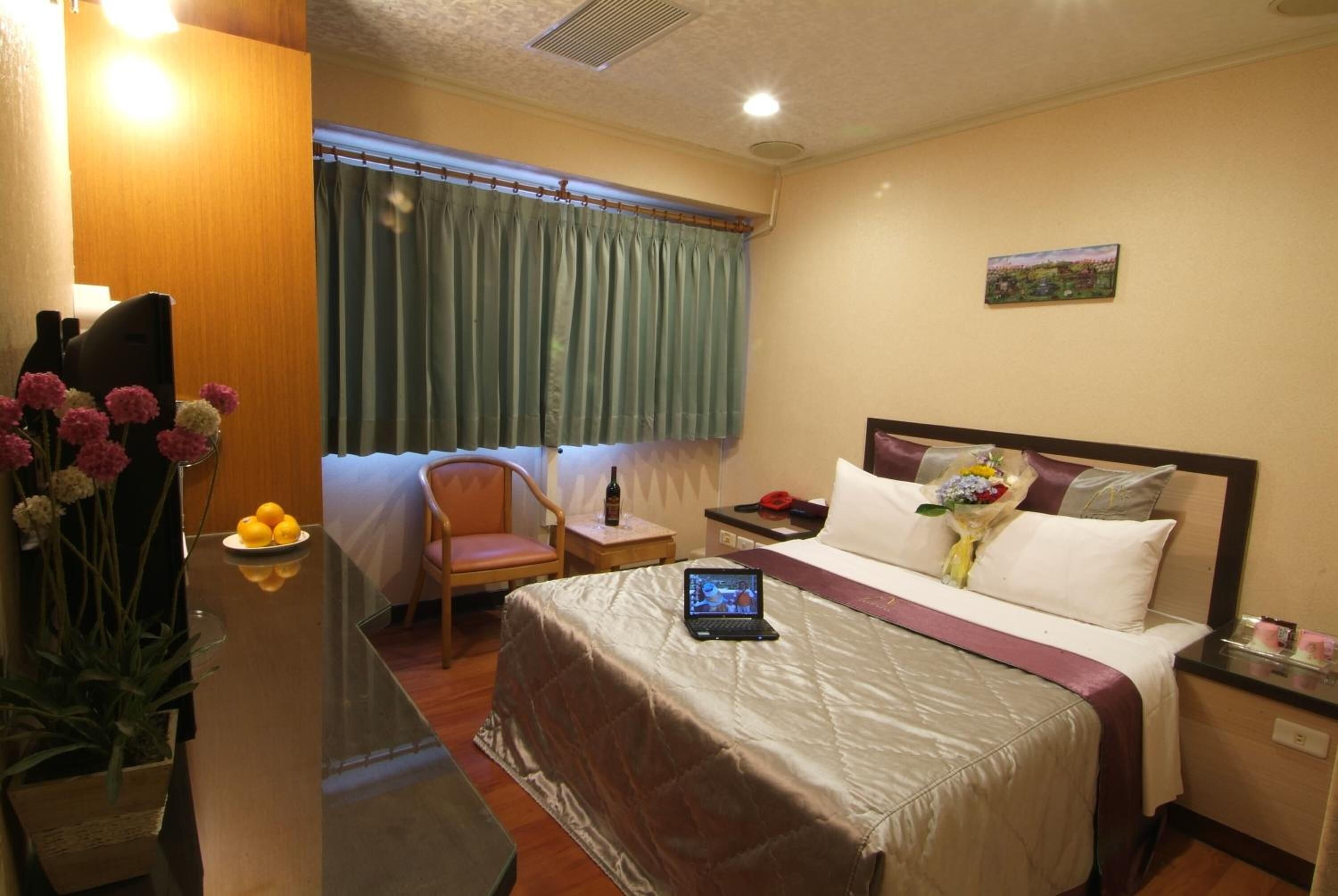 Ein Hotelzimmer mit einem Bett, einem Schreibtisch mit Laptop, einem Stuhl, einem Tisch mit Blumenvase und einem fenster mit Vorhängen.