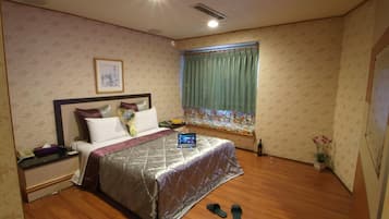 A hotel room with a bed, a bedside table, a laptop, slippers, a window with curtains, and a wall-mounted picture.