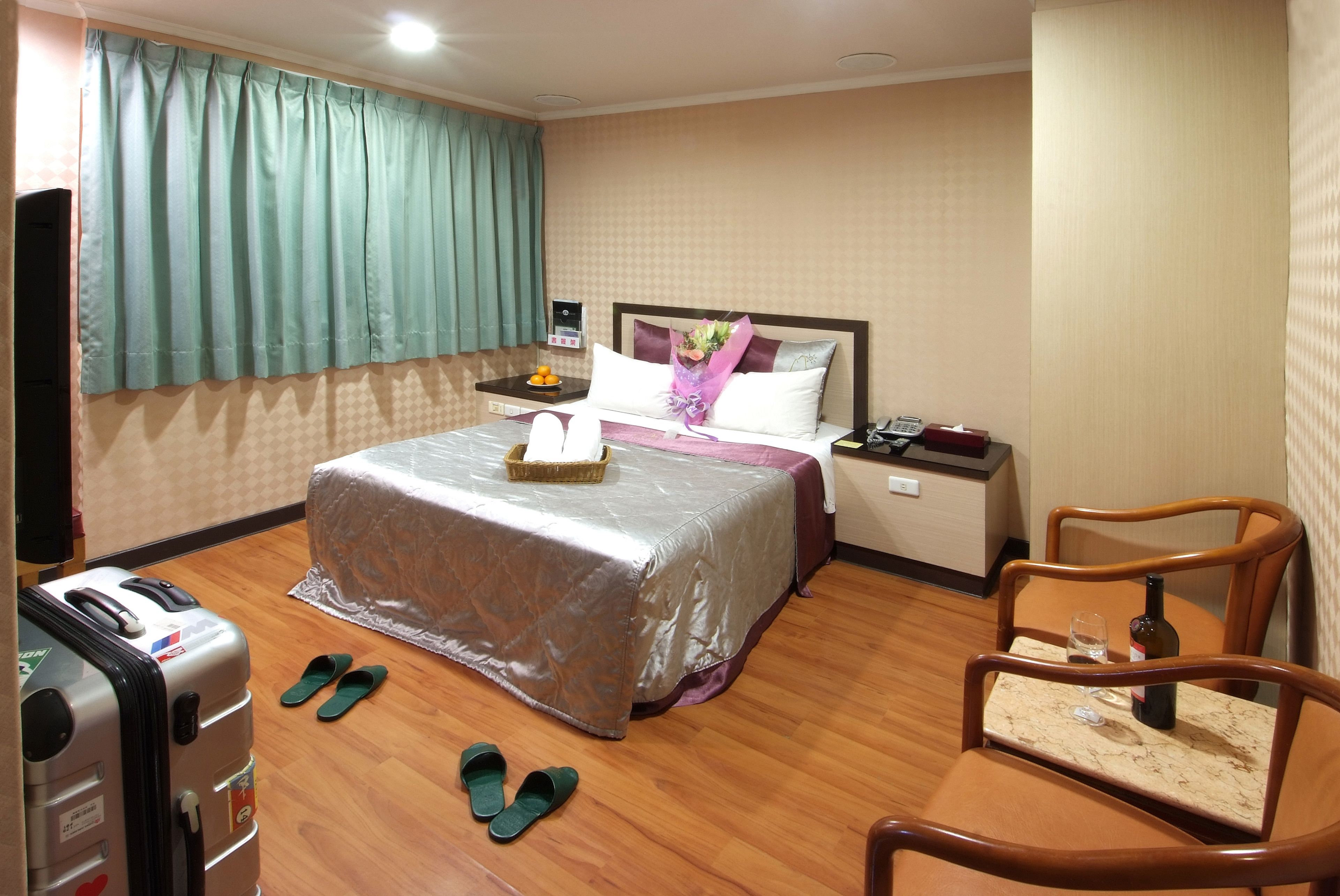 A hotel room with a bed, a desk, a chair, a suitcase, slippers, and a bottle of wine.