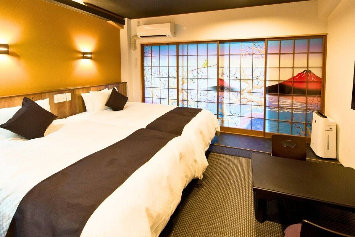 A hotel room with a large bed, two wall-mounted lights, a view through a window with a decorative screen, and a desk with a chair.