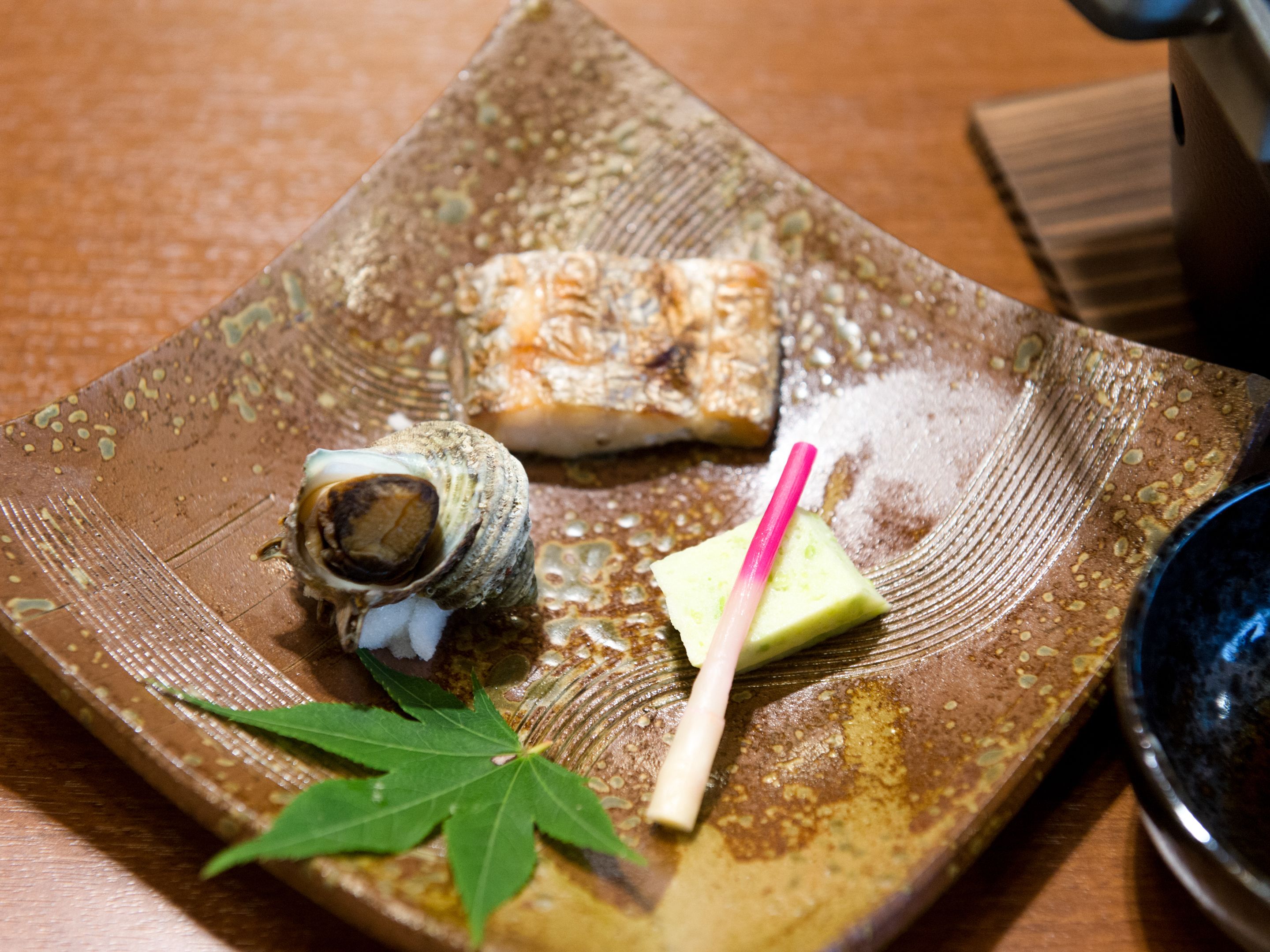 A square ceramic plate with a piece of fish, a green leaf, and a pink straw.