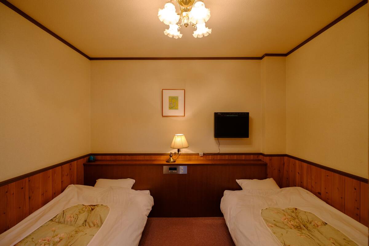 A hotel room with two single beds, a nightstand, a lamp, a flat-screen TV mounted on the wall, and a ceiling light fixture.