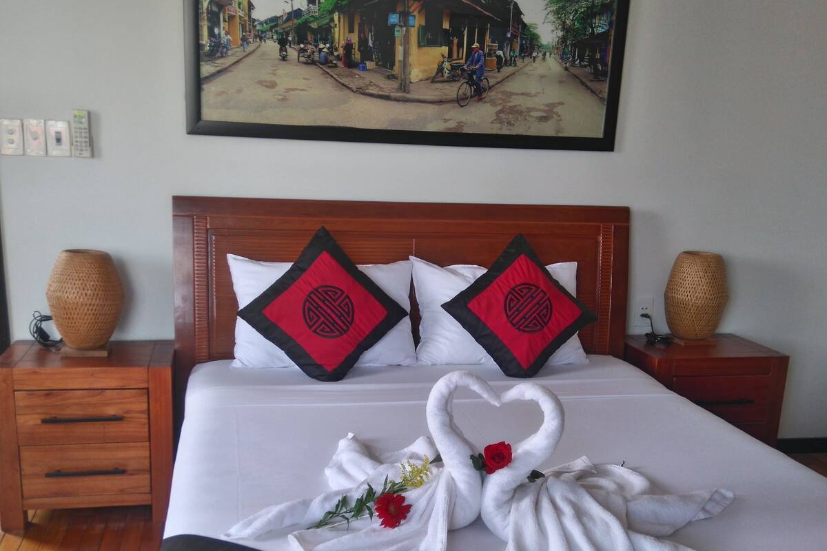 A neatly made bed with a red and black patterned bedspread, two pillows with a red and black design, and a heart-shaped towel arrangement with a red rose.