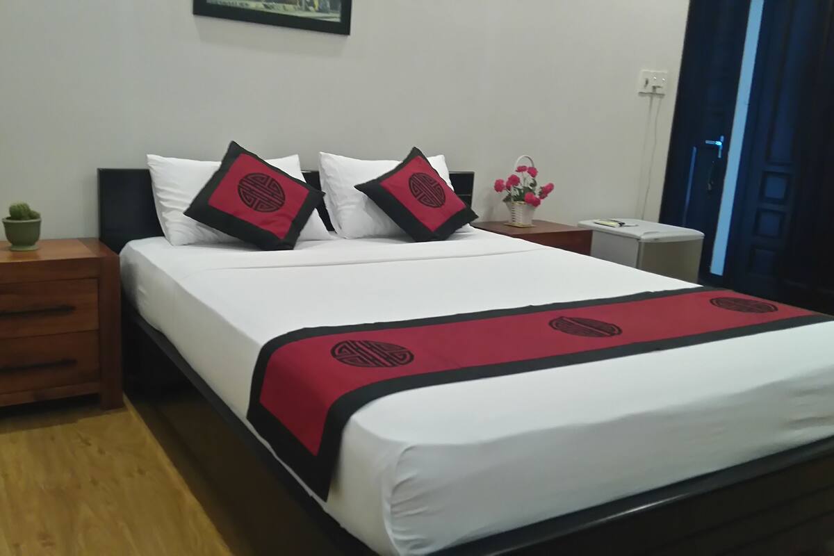 A neatly made bed with white bedding and red and black decorative pillows, a wooden nightstand with a green plant, and a small table with a vase of flowers.