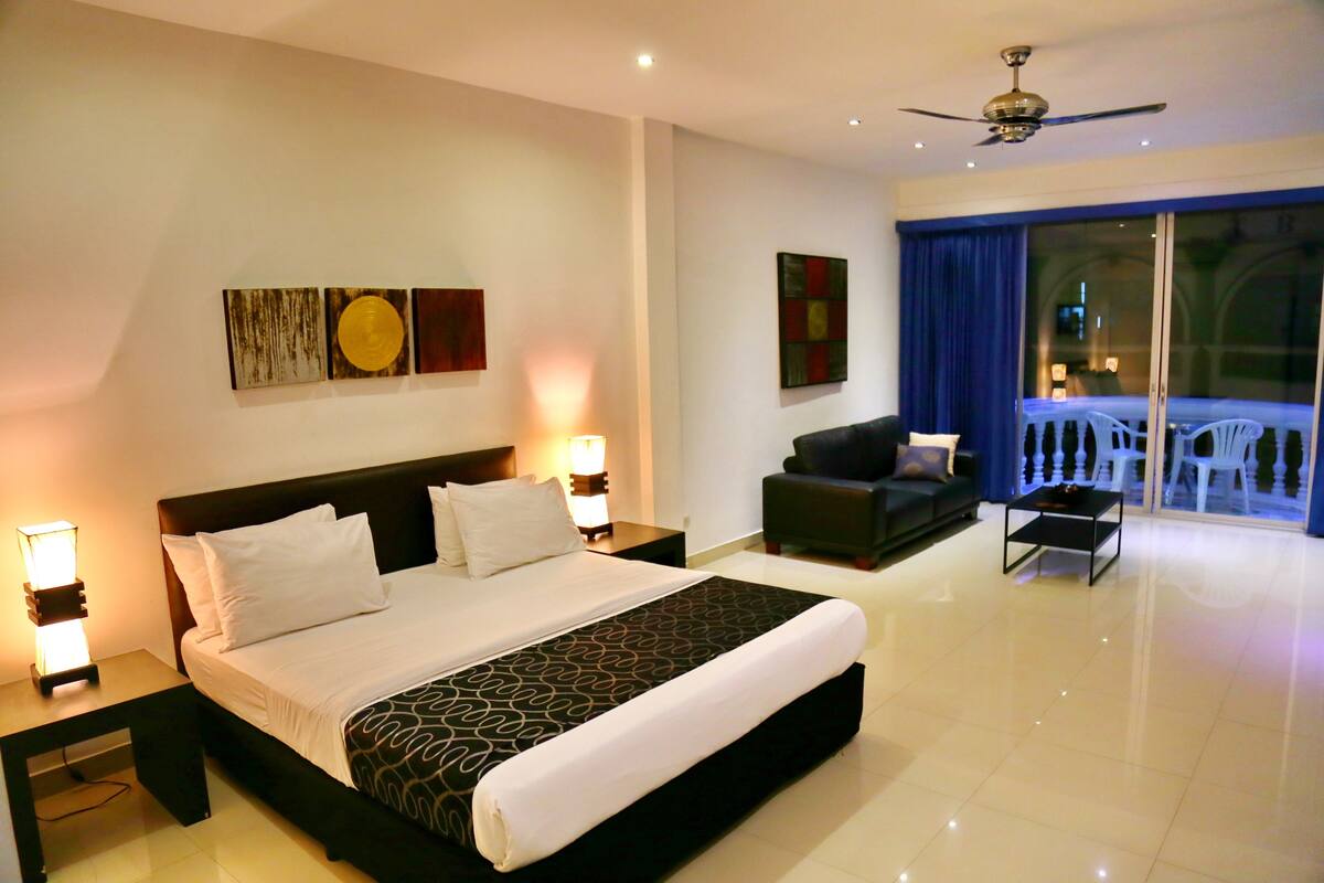 A hotel room with a large bed, two bedside tables, a ceiling fan, and a balcony with white chairs.