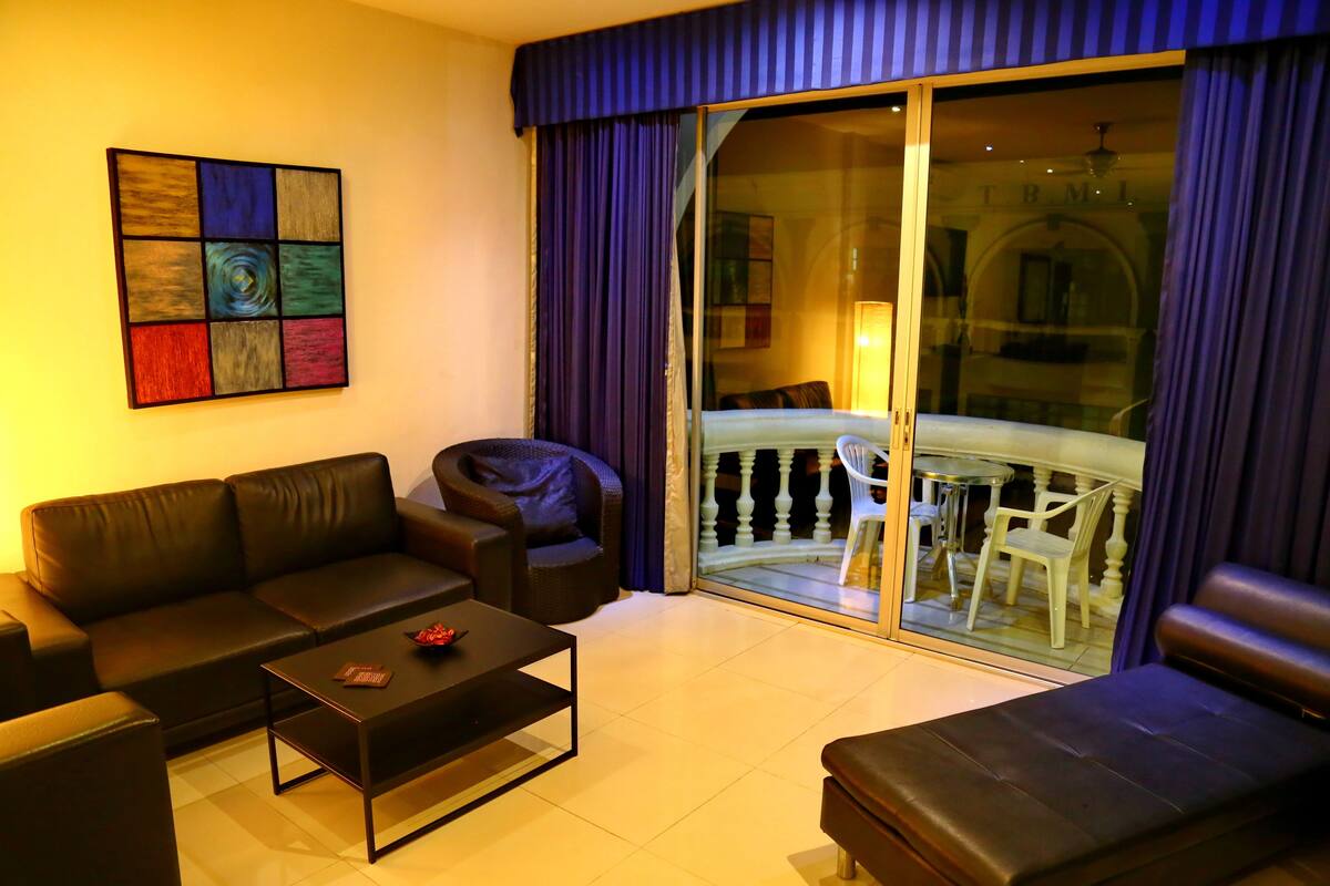 A modern living room with a sofa, a coffee table, and a chair. There is a balcony with a glass door and a view of the outside.
