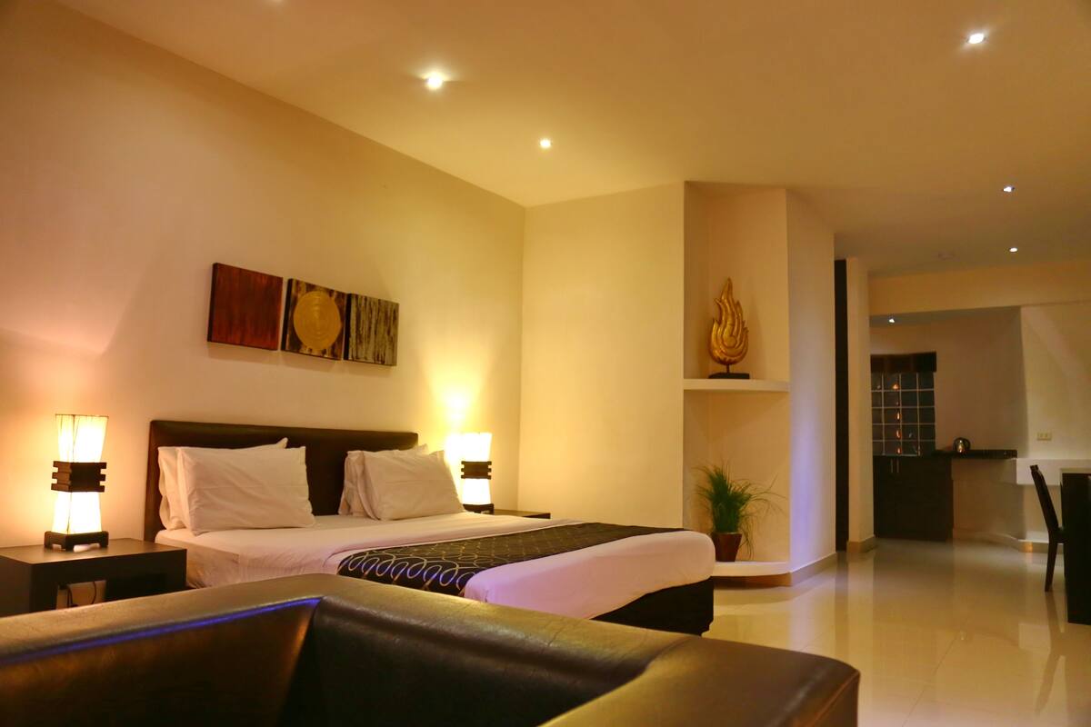 A hotel room with a large bed, bedside lamps, a potted plant, and a view into the kitchen area.