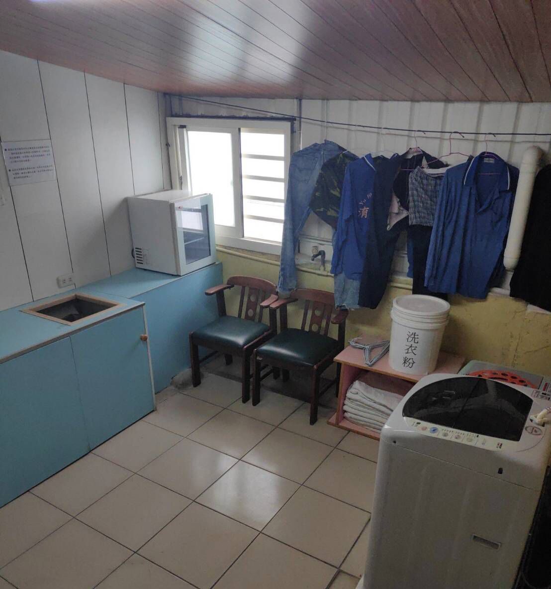 A small, compact kitchen with basic amenities including a microwave, sink, and a washing machine. There are chairs and a table, and clothes hanging on a line.