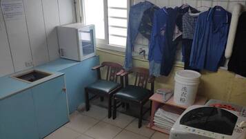A small, compact kitchen with basic amenities including a microwave, sink, and a washing machine. There are chairs and a table, and clothes hanging on a line.