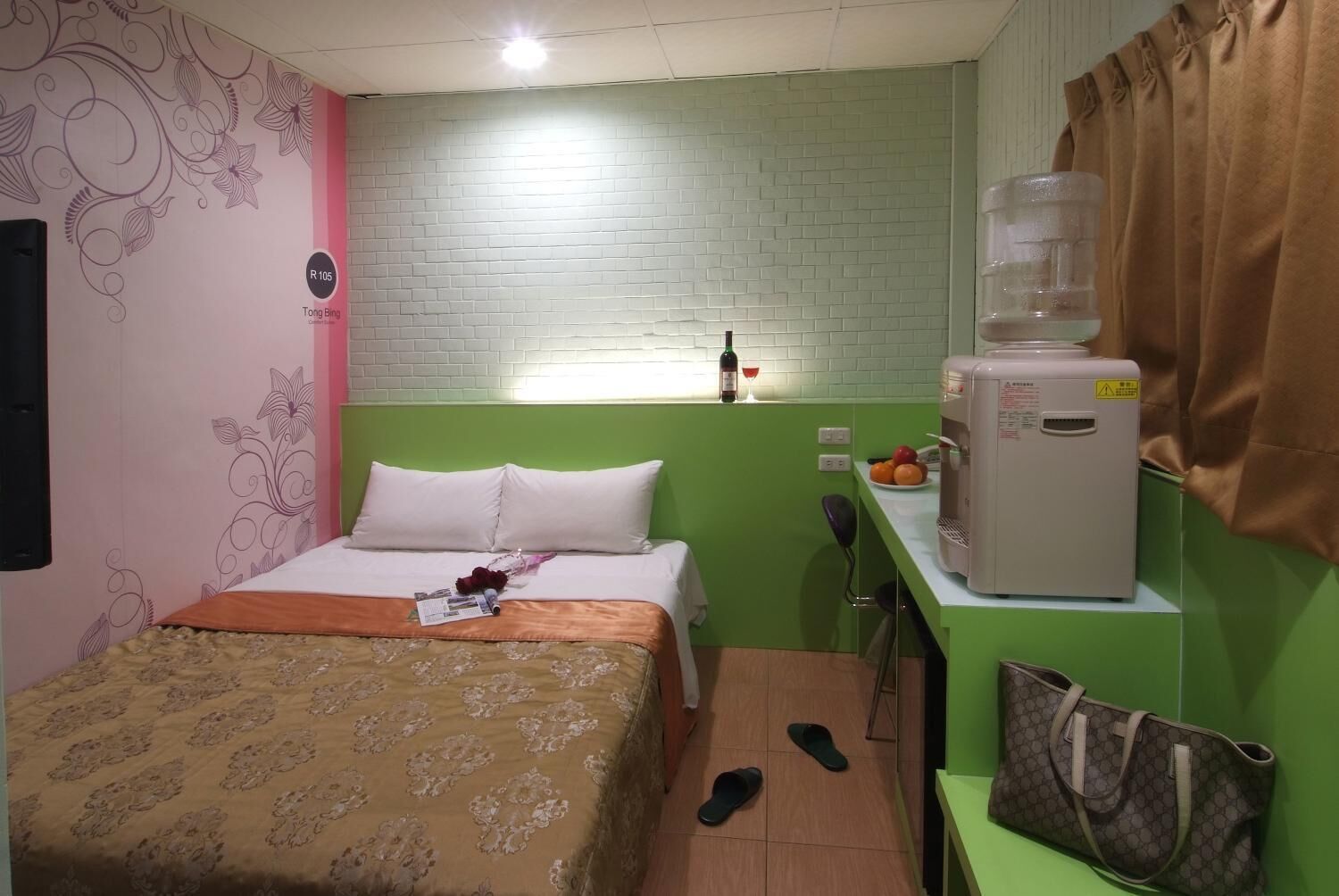 A compact hotel room with a bed, a small kitchenette, a water dispenser, and a green wall.