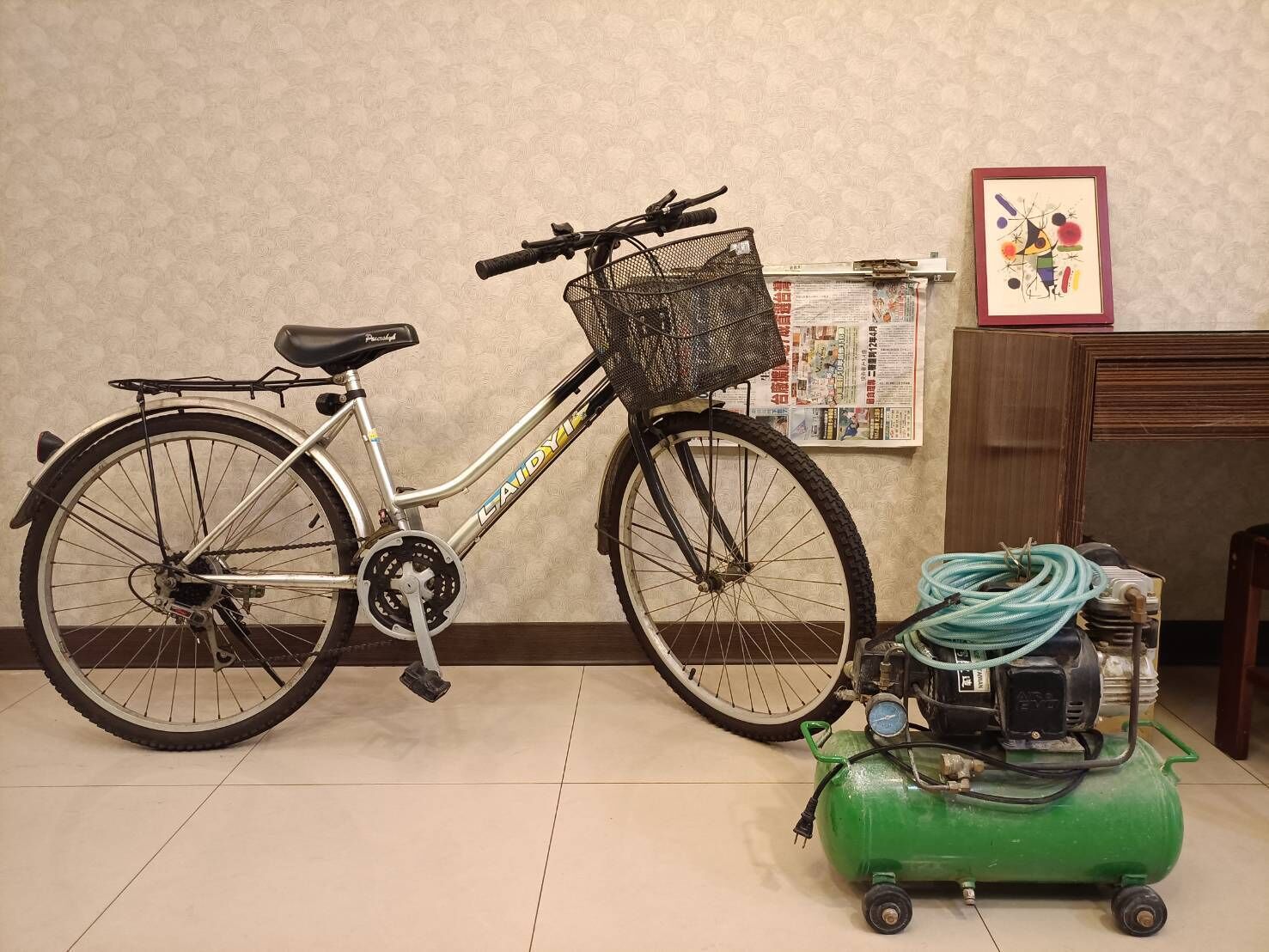 Una bicicleta con una canasta y un compresor de aire verde en una habitación con un tablón de anuncios y un cuadro enmarcado.