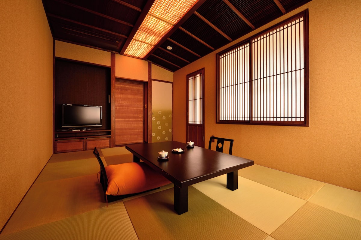 Une pièce traditionnelle japonaise avec un sol recouvert de tatamis, une table basse, un téléviseur, des portes coulissantes et des paravents en papier de riz (shoji).