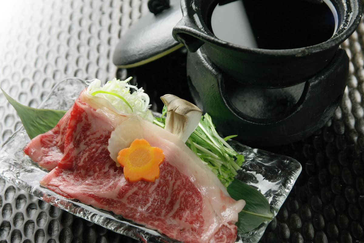 Une assiette en verre transparent contenant de la viande crue, des accompagnements et un pot noir, le tout posé sur une surface texturée.