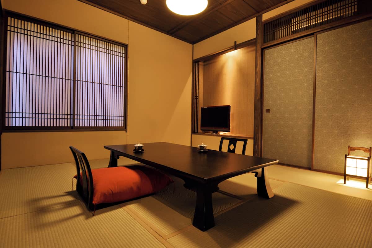 Une pièce de style japonais traditionnel, avec un sol recouvert de tatamis, une table basse, un téléviseur, des portes coulissantes et un coussin rouge.