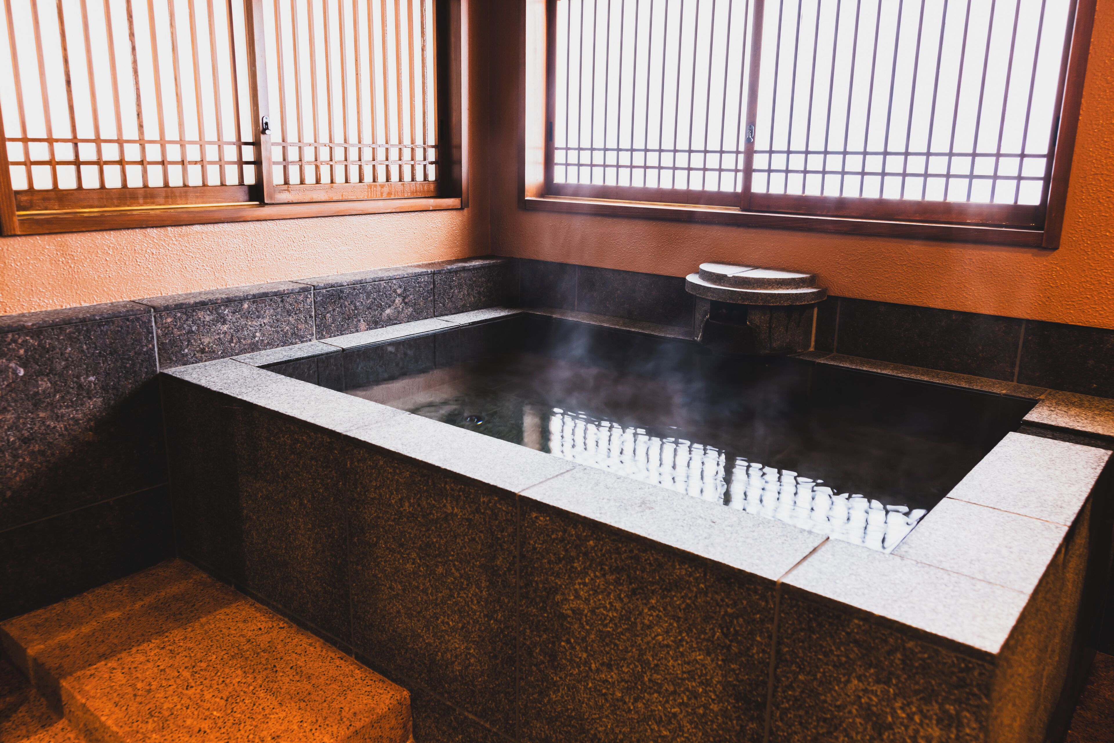 A hot spring bath with a stone edge, a small stool, and a window with vertical bars.