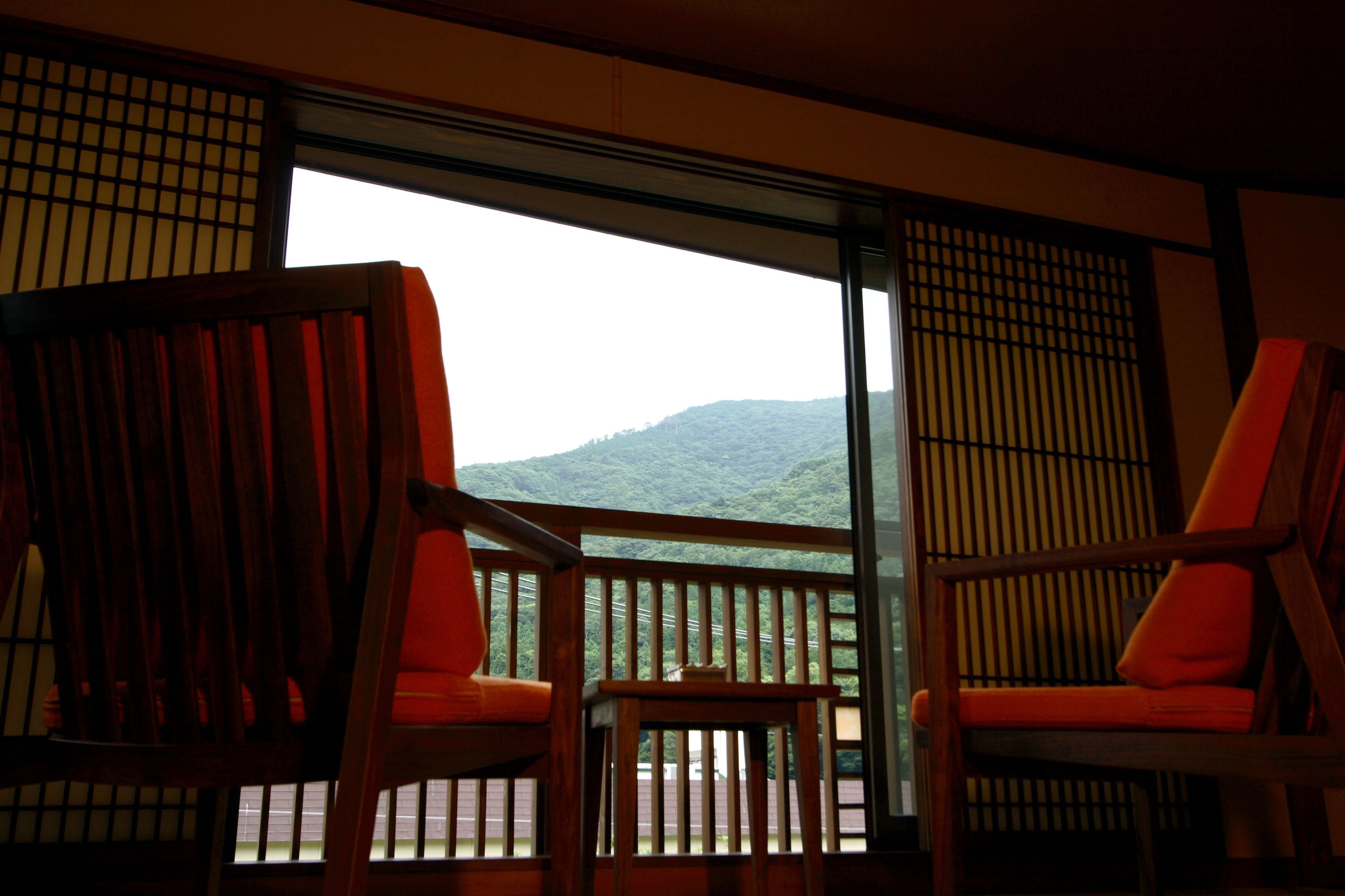 A room with two wooden chairs and a small table, a sliding glass door leading to a balcony with a view of a green landscape.