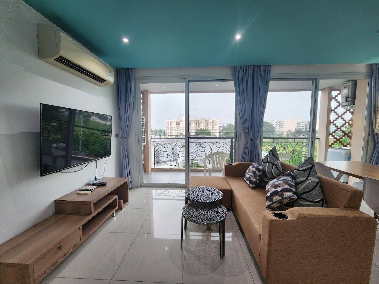 A modern living room with a flat-screen TV, a wooden entertainment unit, a sofa with patterned cushions, and a balcony with a view of buildings.